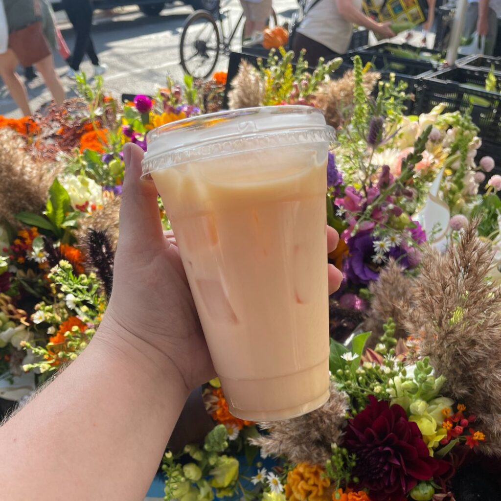 "A hand holding a plastic cup filled with an iced Pumpkin Up Chai Latte in front of a vibrant display of flowers at an outdoor market. The latte has a creamy, orange hue, with the colorful mix of flowers—including purple, orange, and white blossoms—creating a bright and lively background."