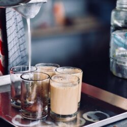 Milky spiced chai being strained through a mesh sieve into glasses on a metal tray