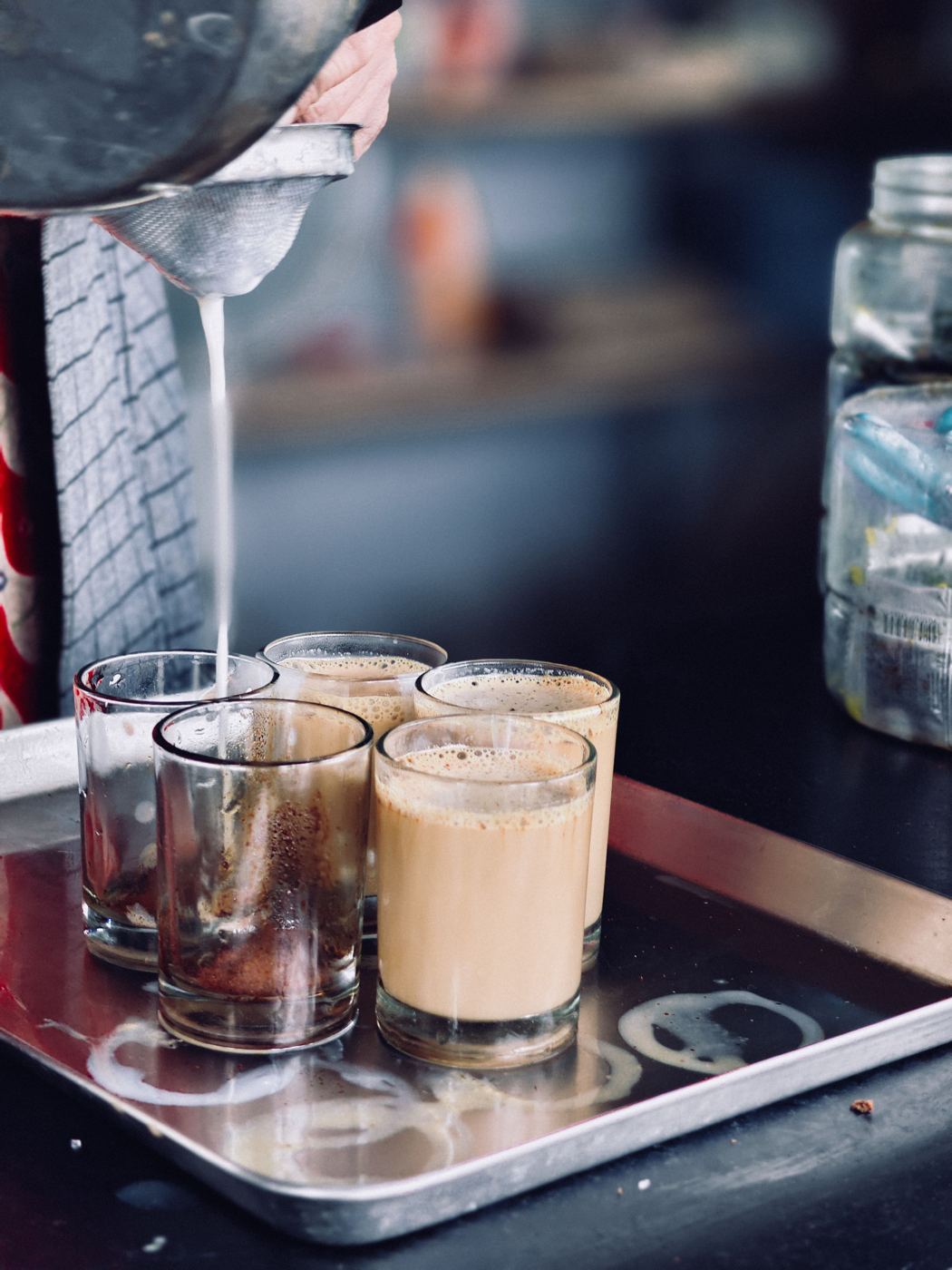 Milky spiced chai being strained through a mesh sieve into glasses on a metal tray