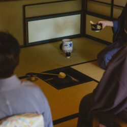 Guests seated on tatami around a sunken charcoal hearth as a host in kimono lifts a lacquered tea container during a formal Japanese tea gathering