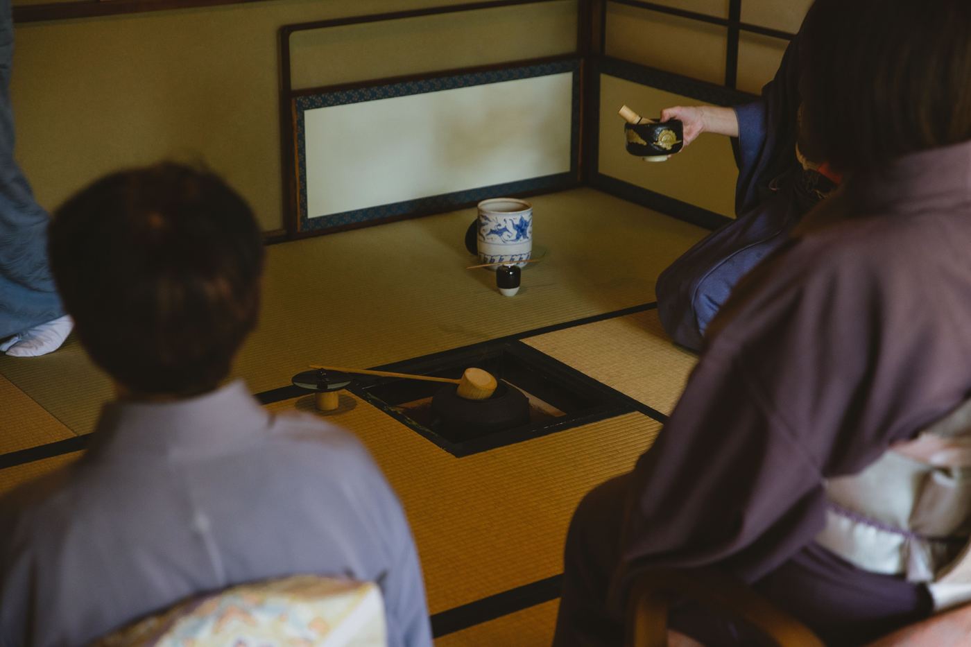 Guests seated on tatami around a sunken charcoal hearth as a host in kimono lifts a lacquered tea container during a formal Japanese tea gathering