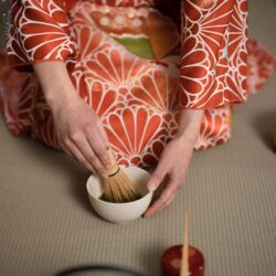 Hands in a kimono sleeve whisking matcha with a bamboo chasen in a ceramic bowl on a tatami mat