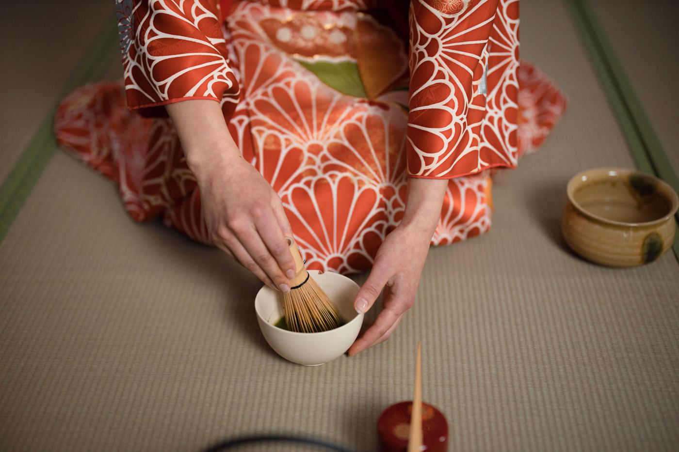 Hands in a kimono sleeve whisking matcha with a bamboo chasen in a ceramic bowl on a tatami mat