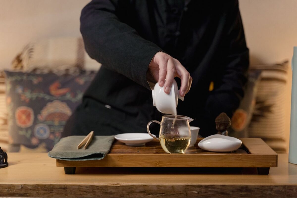 A person in a dark tunic pouring tea from a small gaiwan into a glass pitcher on a wooden tea tray set with cups