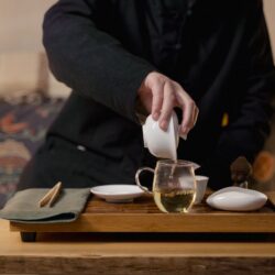 A person in a dark tunic pouring tea from a small gaiwan into a glass pitcher on a wooden tea tray set with cups