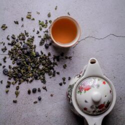 Rolled oolong tea leaves scattered beside a cup of golden brewed tea and a decorative teapot