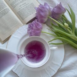 Purple-hued tea poured into a white teacup beside purple tulips and an open book