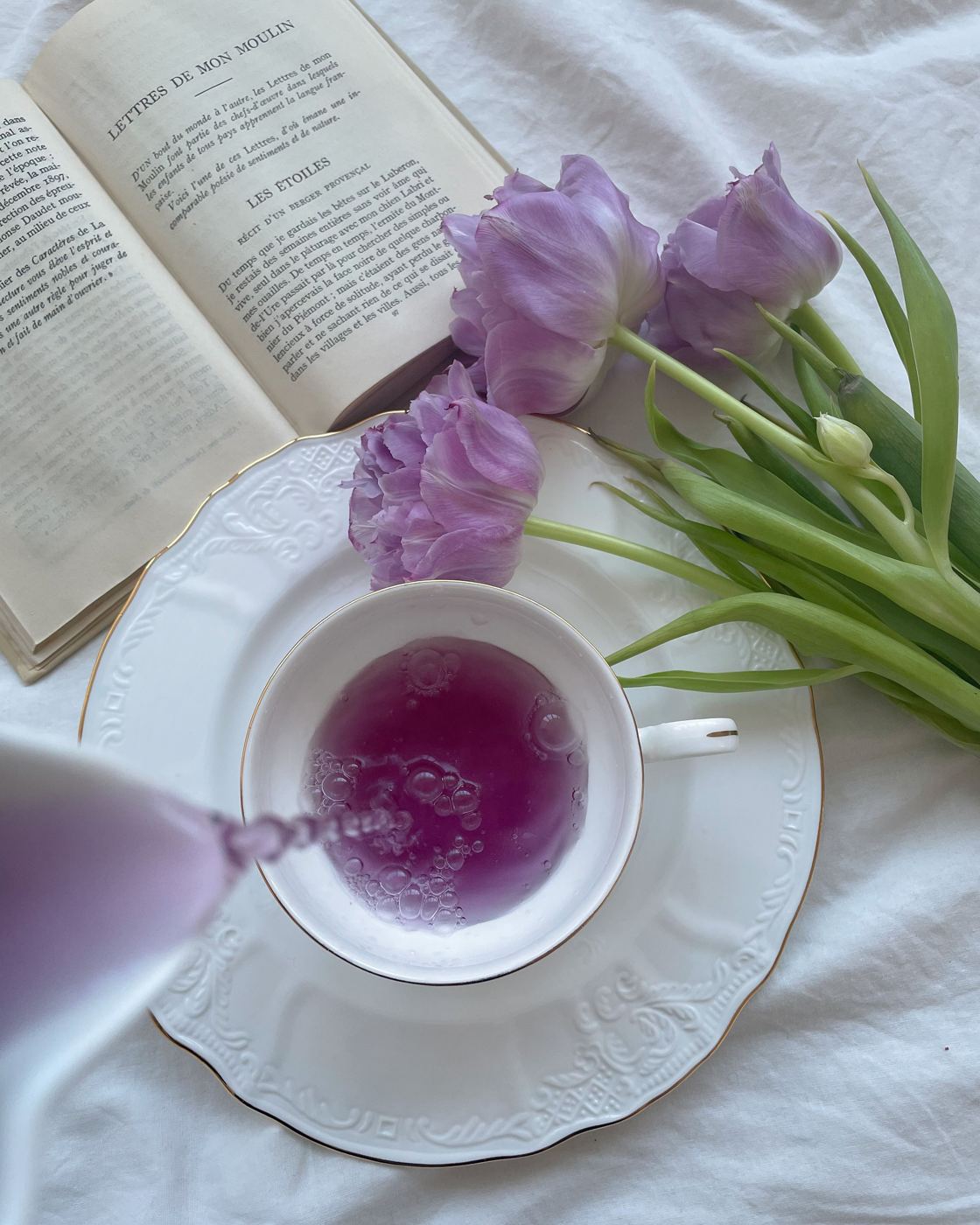 Purple-hued tea poured into a white teacup beside purple tulips and an open book