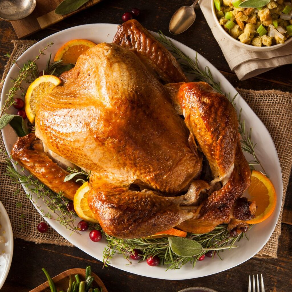 A golden-brown roasted turkey brined with Kumaon Black tea, garnished with orange slices, fresh rosemary, and cranberries. The turkey is plated on a white platter surrounded by other Thanksgiving sides, ready to serve.