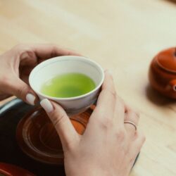 Hands holding a cup of Japanese green tea beside a clay teapot