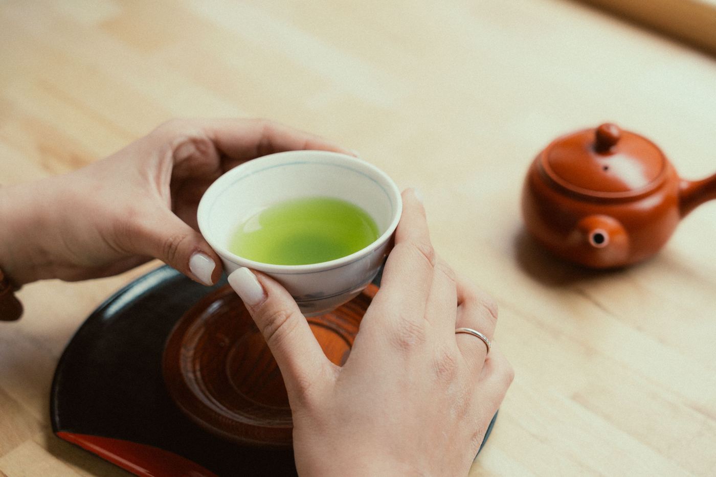 Hands holding a cup of Japanese green tea beside a clay teapot