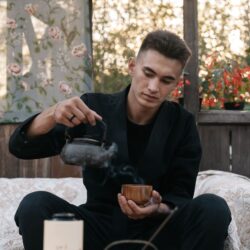 A man pours steaming tea from a dark kettle into a small wooden cup in a cozy, plant-filled room