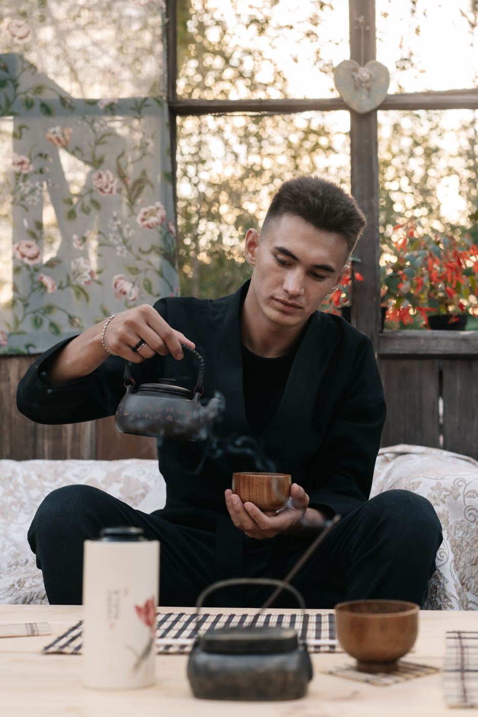 A man pours steaming tea from a dark kettle into a small wooden cup in a cozy, plant-filled room