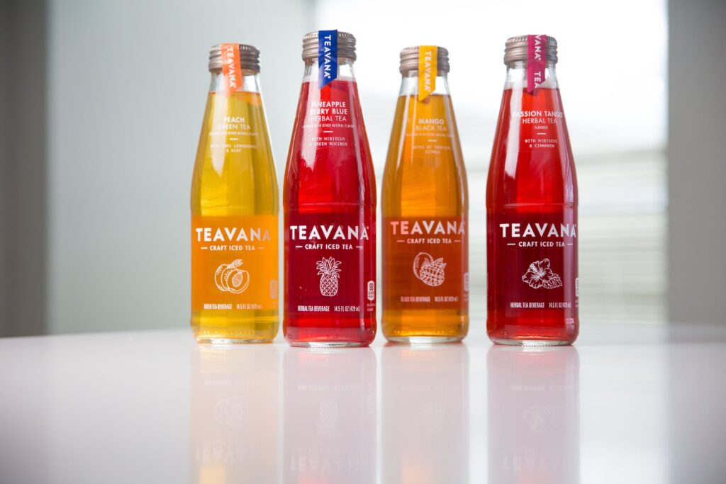 Four bottles of Teavana Craft Iced Tea are displayed on a reflective white surface. The flavors include Peach Green Tea, Pineapple Berry Blue Herbal Tea, Mango Black Tea, and Passion Tango Herbal Tea. Each bottle features vibrant colors representing its flavor, clear labels, and unique fruit or botanical illustrations. The teas are packaged in sleek, transparent bottles with metallic caps and color-coded tags.