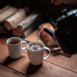 Tea being poured from a dark teapot into two white mugs on a rustic wooden table