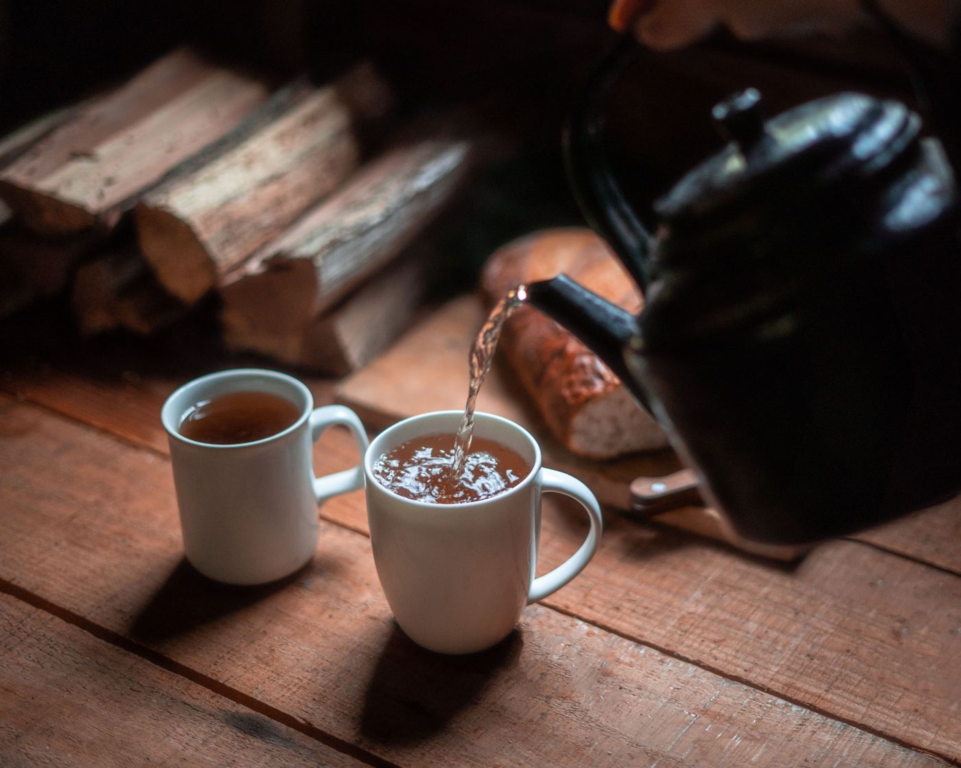 Tea being poured from a dark teapot into two white mugs on a rustic wooden table