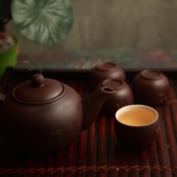 A dark purple clay Yixing teapot with matching small cups on a bamboo tray, steeped tea poured into one cup