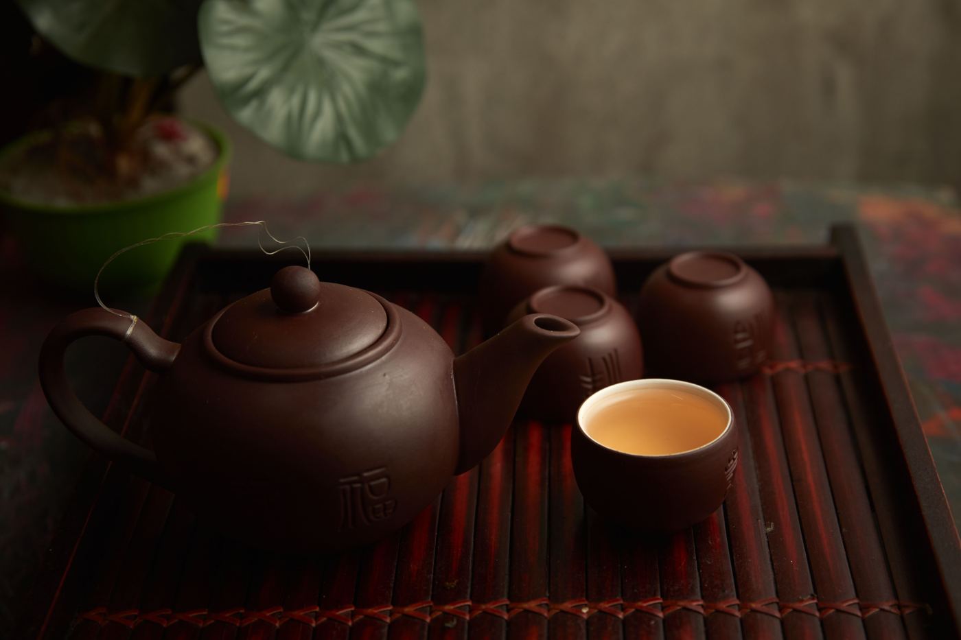 A dark purple clay Yixing teapot with matching small cups on a bamboo tray, steeped tea poured into one cup