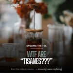 A stylish French press filled with an herbal tisane surrounded by glassware, with blurred orange flowers in the background. The text overlay includes “MOODY TEA NEWS” at the top, “Spilling the Tea” in smaller text, and “WTF Are ‘Tisanes???’” in bold letters. At the bottom, it says: “For the latest news → moodyteas.co/blog