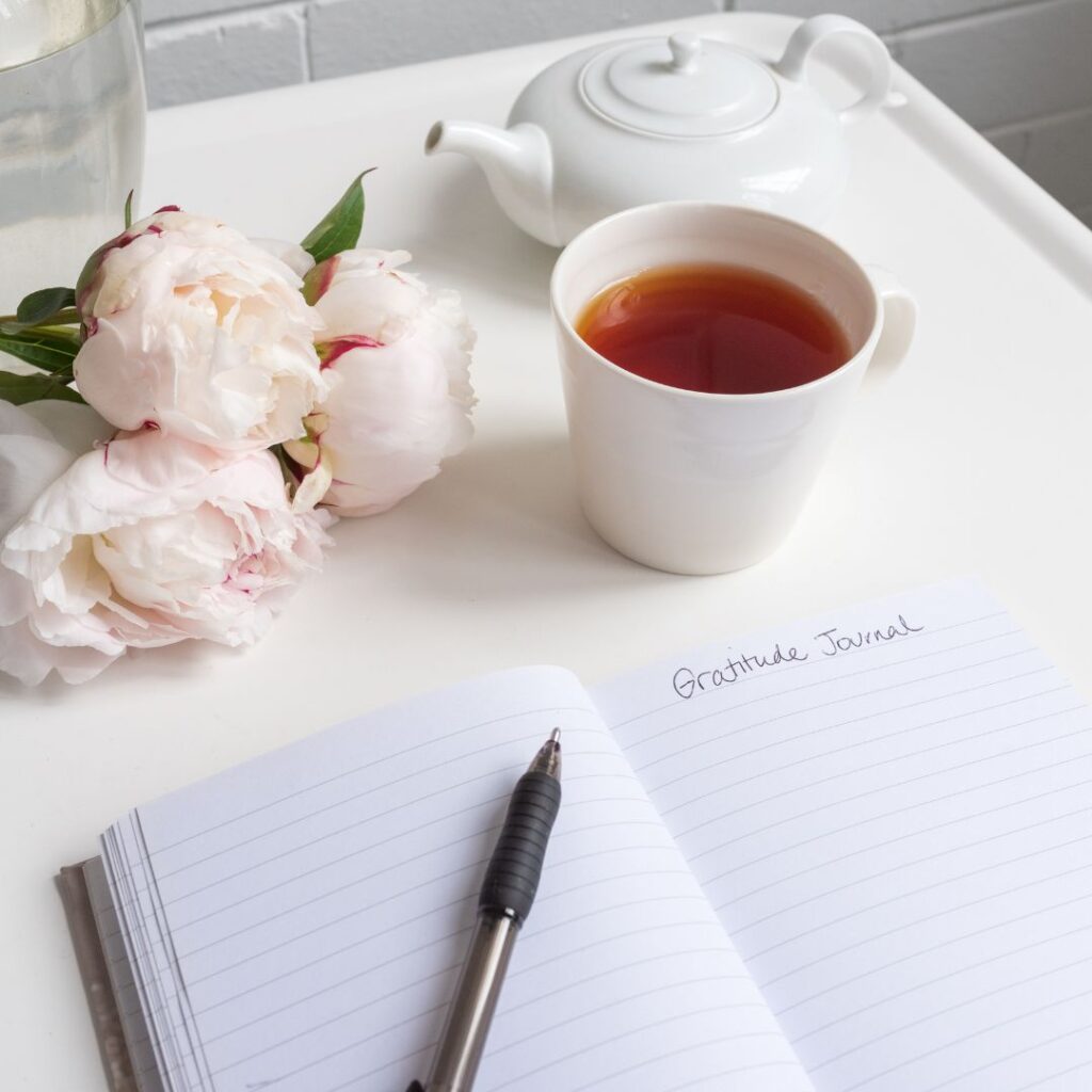 A serene setup of a gratitude journal open on a white table, accompanied by a cup of tea in a white mug, a white teapot, and soft pink peonies. The journal has "Gratitude Journal" written at the top of the page, creating a peaceful and reflective scene.