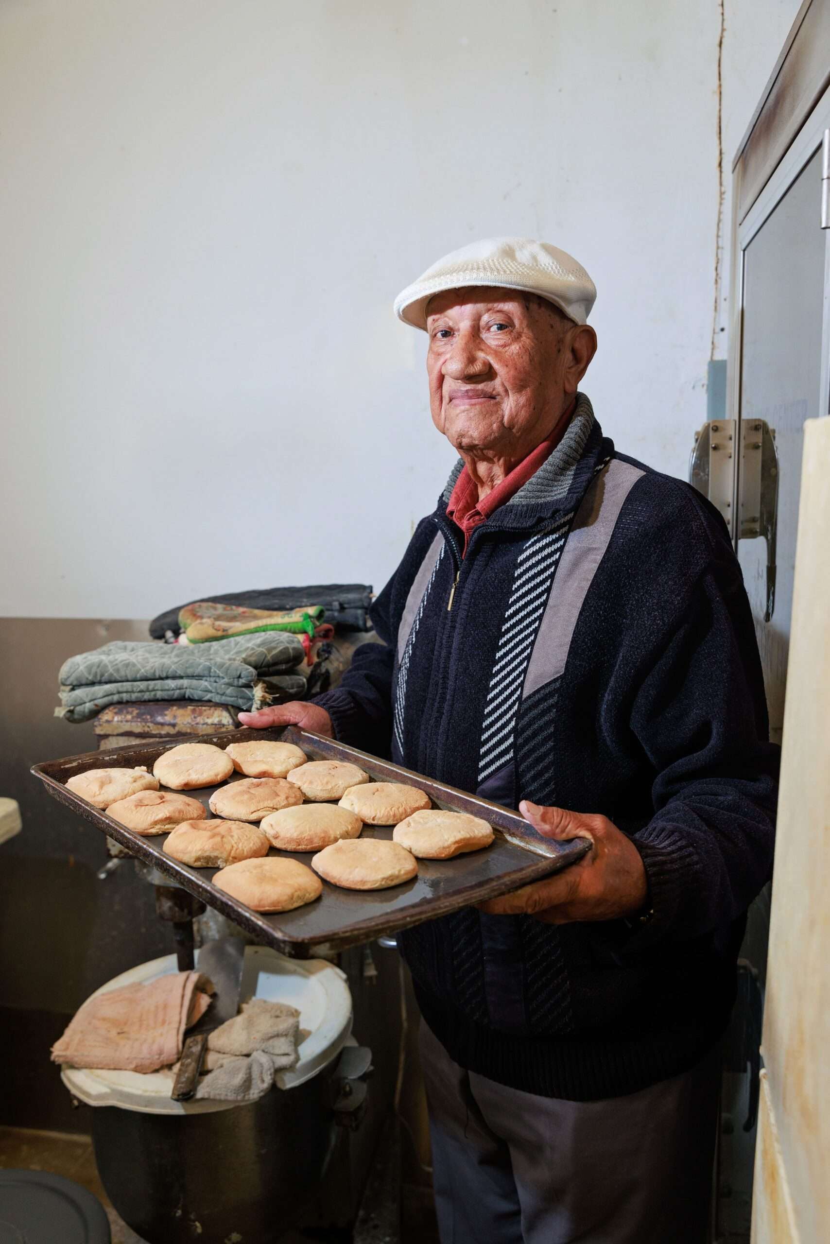 An elderly African American man, Junius Dillard, wearing a white flat cap and dark sweater, holds a baking tray with freshly baked tea cakes. The background features a simple kitchen setting. The image highlights the tradition of tea cake baking in African American culture.