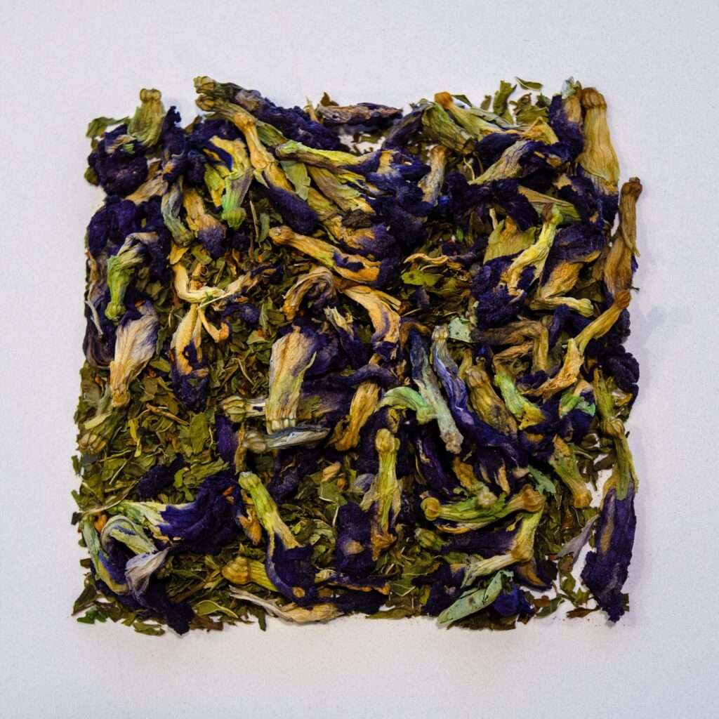 A close-up of loose leaf Transcendence tea blend by Moody Teas, featuring a vibrant mix of deep blue butterfly pea flowers, green spearmint leaves, and fine rooibos, lightly infused with vanilla essence on a white background.
