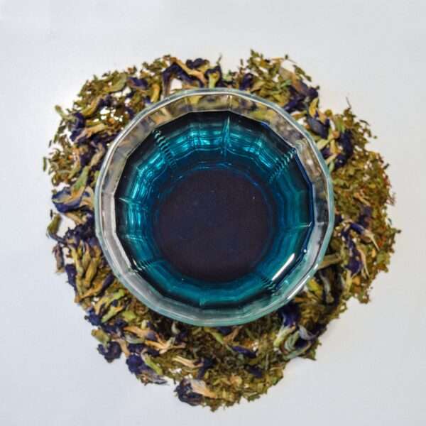 A glass teacup filled with vibrant blue tea sits centered on a white background, surrounded by a circular arrangement of loose leaf rooibos, spearmint, and butterfly pea flowers, essenced with vanilla.