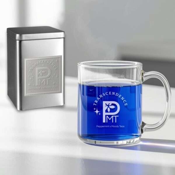 A glass mug of vibrant blue Transcendence tea sits on a white surface beside a silver embossed tin, both featuring the Transcendence logo for the Peppermint x Moody Teas collaboration. Steam rises gently from the mug, highlighting the tea’s striking color and elegant presentation.
