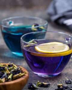 Photo of two glass cups filled with butterfly pea flower tea, one deep blue and the other shifting to purple with a slice of lemon resting on top. Loose dried butterfly pea flowers sit in a wooden bowl nearby. The image showcases the natural pH-based color change reaction caused by adding citrus.