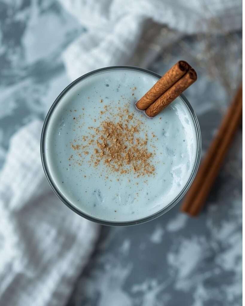A close-up overhead shot of a creamy icy-blue coquito in a glass, sprinkled with ground cinnamon and garnished with two cinnamon sticks. The background features a soft marble surface and a white textured cloth.