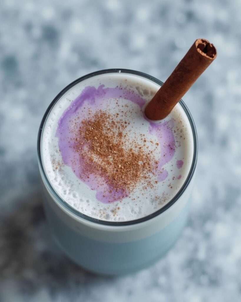 A glass of icy-blue coquito with a lavender swirl on the surface, sprinkled with cinnamon and garnished with a single cinnamon stick. The background is a soft, cool-toned marble texture.