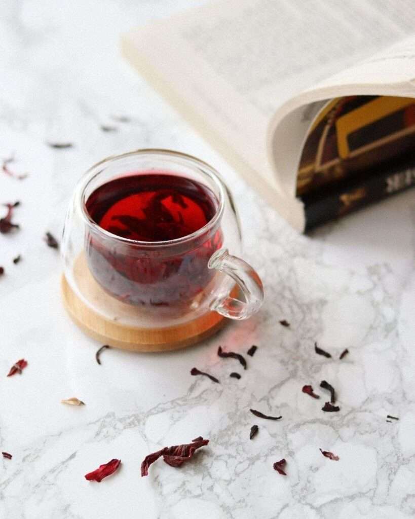 Glass mug of deep red herbal tea on a marble surface beside an open book, representing a slow, intentional tea routine.