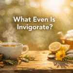 Warm, sunlit tea scene with a steaming white cup of tea on a rustic wooden table, surrounded by loose tea leaves, rosemary sprigs, sliced lemon, and a small yellow flower. Soft golden light and blurred greenery fill the background, with the text “What Even Is Invigorate?” centered above the tea.