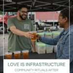 A candid photo at an outdoor farmers market shows John, a bearded Black man wearing glasses and a green Moody Teas t-shirt, smiling as he hands a clear plastic cup of iced tea to a woman in a denim jacket across a black-covered table. Three additional cups of iced tea sit on the table in front of clear ingredient bins. A black canopy tent and a red truck are visible in the background. Overlaid text reads, “Love Is Infrastructure: Community Rituals After Valentine’s Day.”