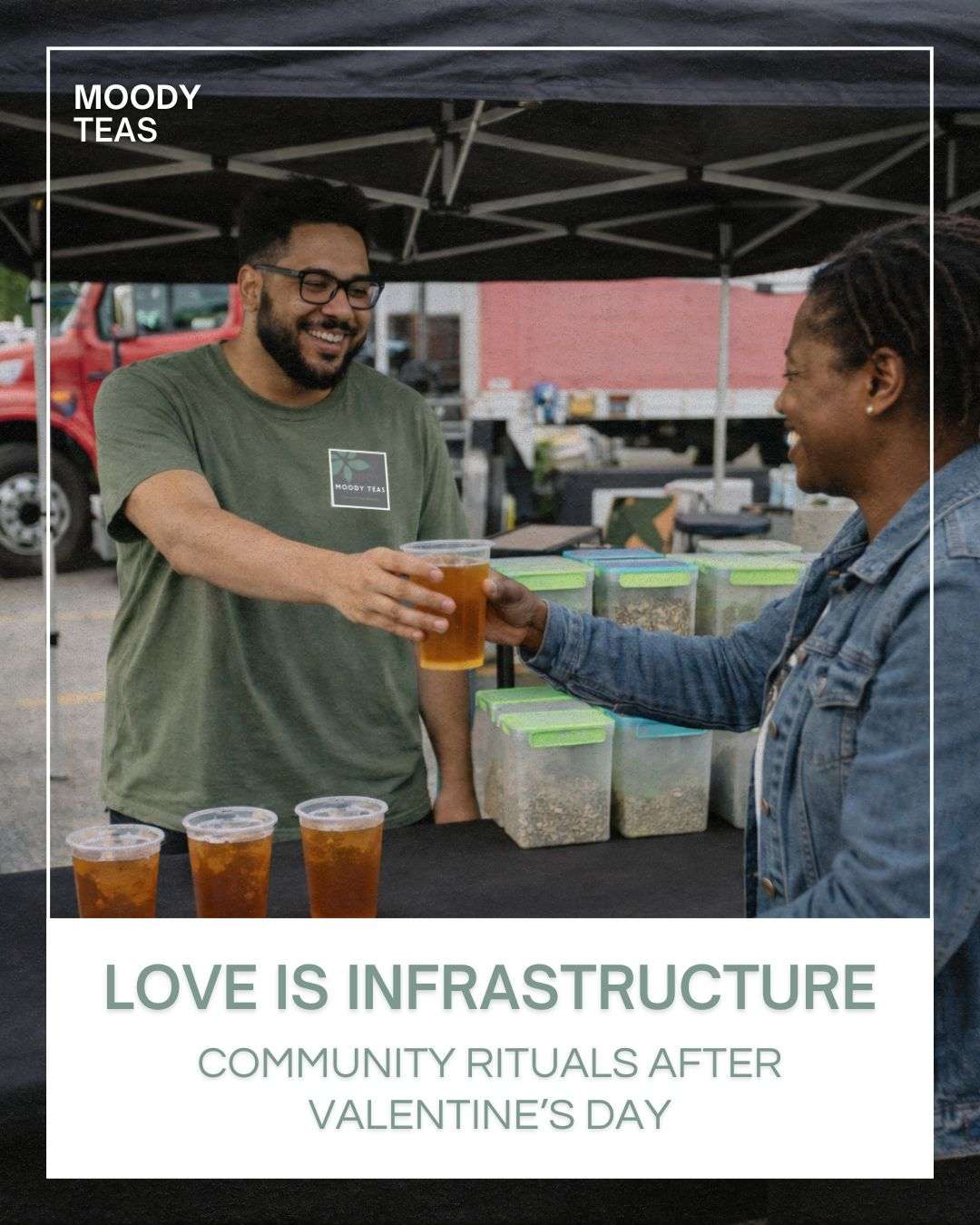 A candid photo at an outdoor farmers market shows John, a bearded Black man wearing glasses and a green Moody Teas t-shirt, smiling as he hands a clear plastic cup of iced tea to a woman in a denim jacket across a black-covered table. Three additional cups of iced tea sit on the table in front of clear ingredient bins. A black canopy tent and a red truck are visible in the background. Overlaid text reads, “Love Is Infrastructure: Community Rituals After Valentine’s Day.”